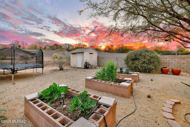 yard at dusk featuring a garden, a fenced backyard, an outdoor structure, a storage unit, and a trampoline