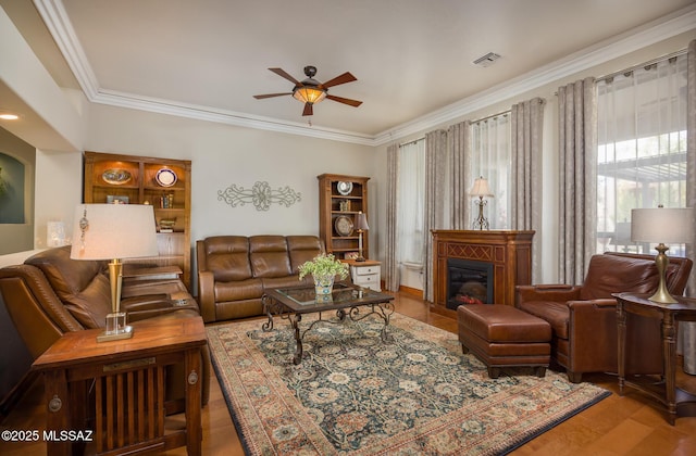 living room with visible vents, ornamental molding, a ceiling fan, a glass covered fireplace, and wood finished floors