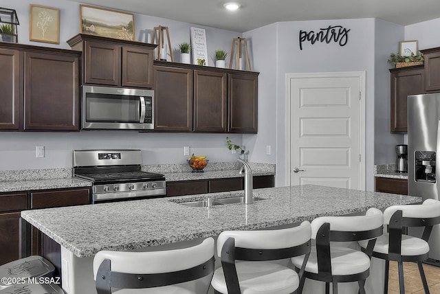 kitchen featuring dark brown cabinets, light stone countertops, a breakfast bar, stainless steel appliances, and a sink