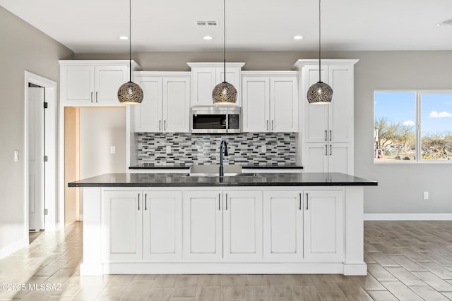 kitchen featuring dark countertops, a kitchen island with sink, a sink, stainless steel microwave, and tasteful backsplash