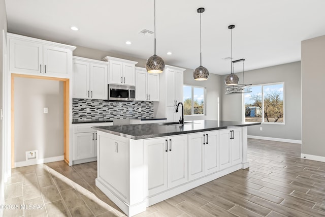 kitchen with a center island with sink, stainless steel microwave, dark countertops, decorative backsplash, and wood tiled floor