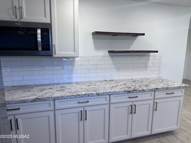 kitchen with light wood-type flooring, open shelves, stainless steel microwave, light stone counters, and backsplash