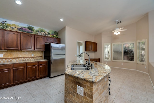 kitchen featuring light stone counters, a ceiling fan, an island with sink, freestanding refrigerator, and a sink