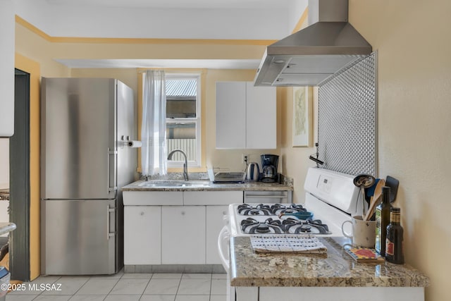 kitchen with a sink, light stone counters, white cabinetry, freestanding refrigerator, and wall chimney range hood