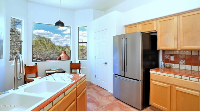 kitchen featuring tile countertops, light brown cabinets, freestanding refrigerator, a sink, and decorative light fixtures