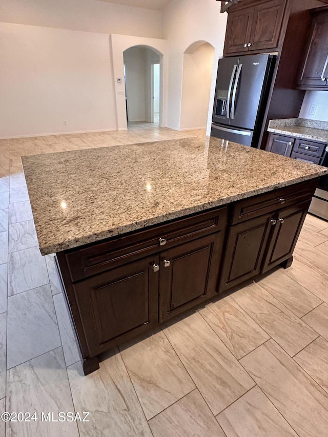 kitchen with a kitchen island, light stone countertops, dark brown cabinetry, stainless steel fridge with ice dispenser, and arched walkways