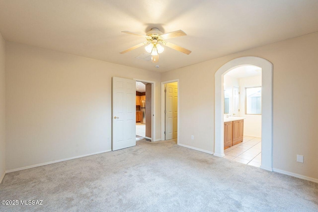 unfurnished bedroom featuring ensuite bathroom, a ceiling fan, arched walkways, baseboards, and light colored carpet