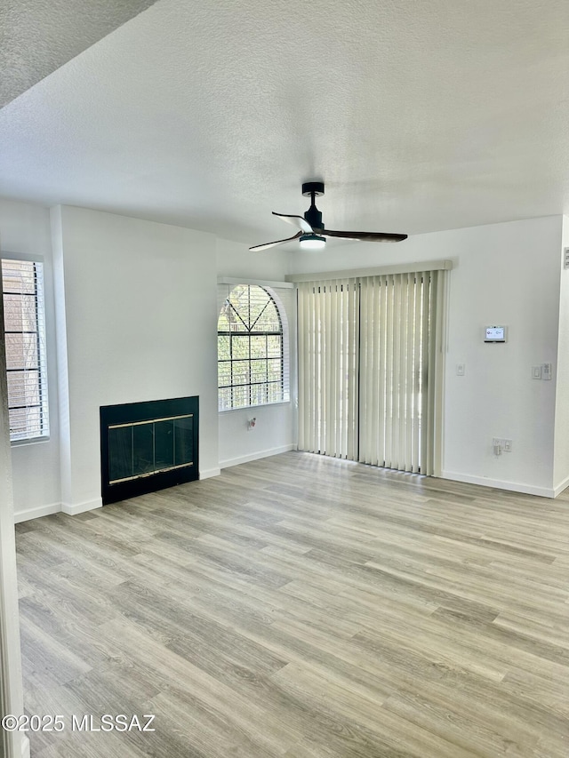 unfurnished living room with baseboards, ceiling fan, light wood-type flooring, a glass covered fireplace, and a textured ceiling