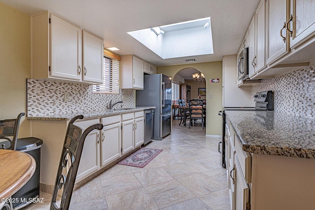 kitchen featuring stone counters, arched walkways, a sink, stainless steel appliances, and tasteful backsplash