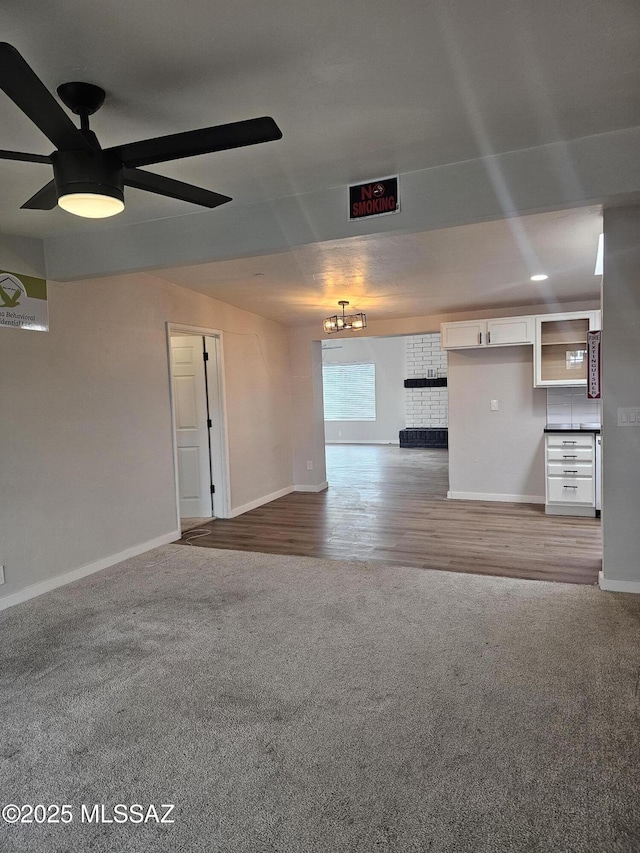 unfurnished living room featuring a brick fireplace, ceiling fan with notable chandelier, carpet, and baseboards