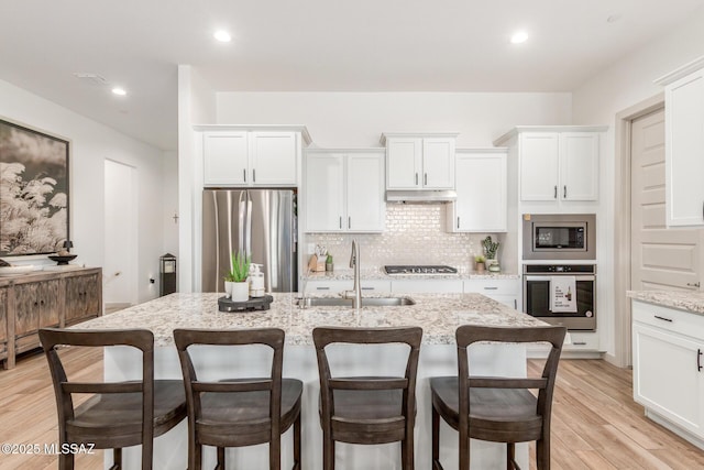 kitchen with light wood-style flooring, a sink, stainless steel appliances, under cabinet range hood, and backsplash