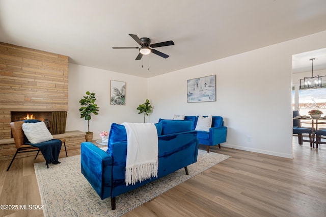 living area with a stone fireplace, ceiling fan with notable chandelier, baseboards, and wood finished floors