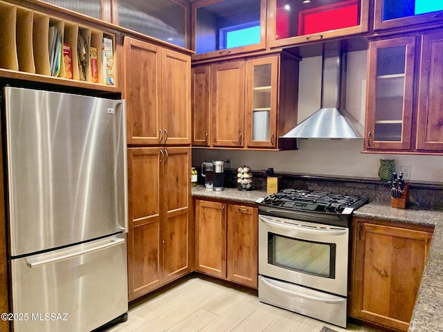 kitchen with glass insert cabinets, brown cabinets, appliances with stainless steel finishes, and wall chimney range hood