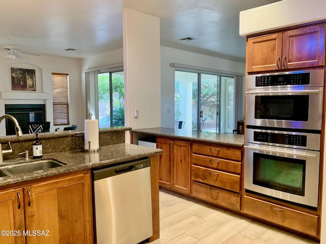 kitchen featuring a sink, a glass covered fireplace, stainless steel appliances, dark stone counters, and brown cabinetry