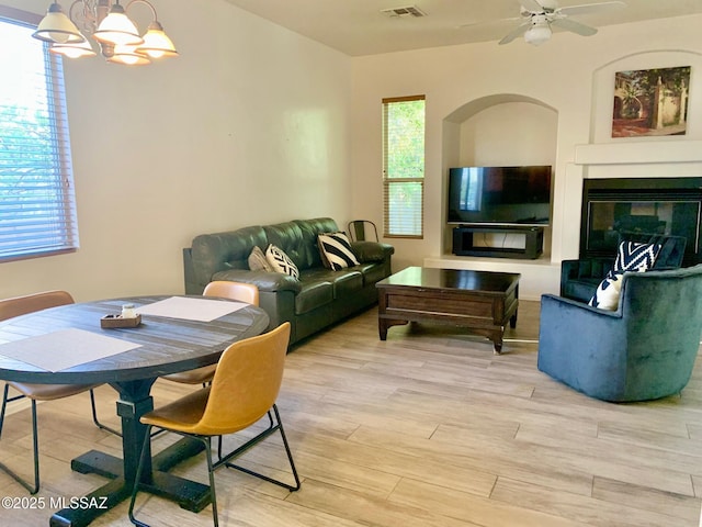 living area with light wood finished floors, a glass covered fireplace, ceiling fan with notable chandelier, and visible vents