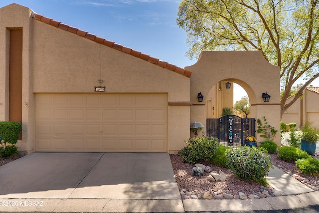 pueblo revival-style home with an attached garage, a tiled roof, stucco siding, driveway, and a gate