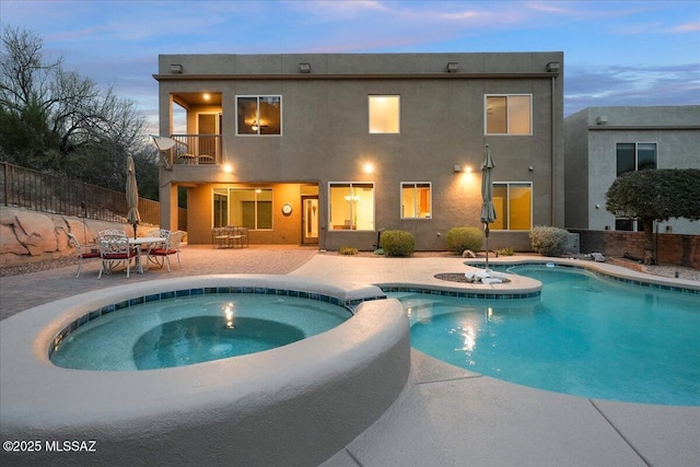 back of house at dusk with a pool with connected hot tub, stucco siding, a patio area, fence, and a balcony