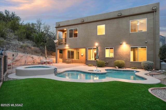 back of house at dusk featuring an in ground hot tub, stucco siding, a lawn, and a balcony