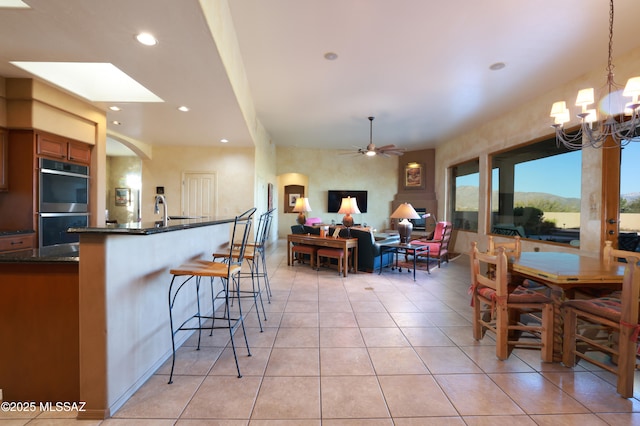 kitchen with a breakfast bar area, light tile patterned floors, brown cabinetry, double oven, and ceiling fan with notable chandelier