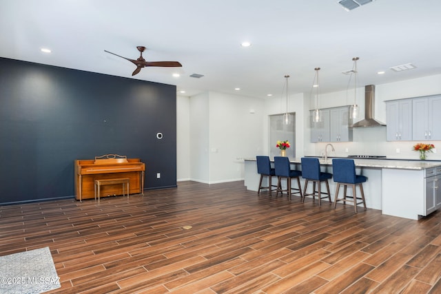 kitchen with ceiling fan, a kitchen island with sink, wall chimney exhaust hood, a kitchen bar, and dark wood-style floors