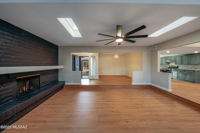 unfurnished living room with visible vents, light wood-style flooring, a ceiling fan, a skylight, and a brick fireplace