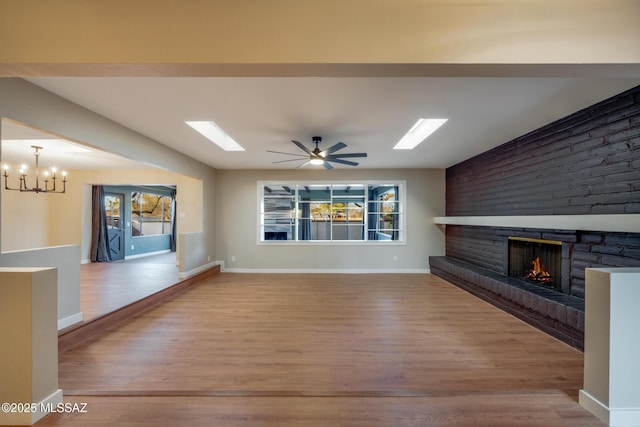 unfurnished living room with a brick fireplace, baseboards, ceiling fan with notable chandelier, a skylight, and wood finished floors