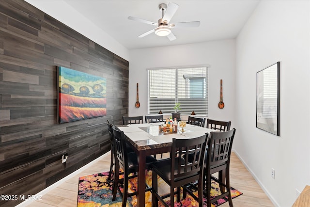 dining room featuring a ceiling fan, wood walls, light wood finished floors, baseboards, and an accent wall