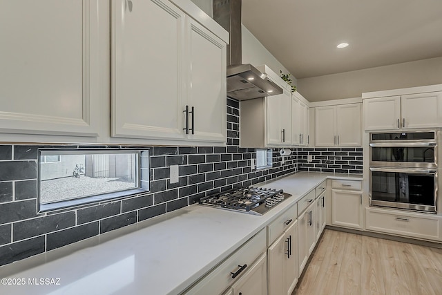 kitchen with decorative backsplash, light countertops, white cabinets, appliances with stainless steel finishes, and wall chimney exhaust hood