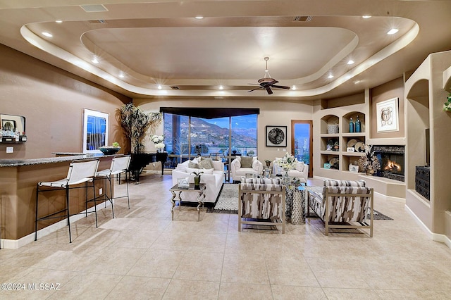 tiled living area with built in features, a ceiling fan, visible vents, a tray ceiling, and a warm lit fireplace