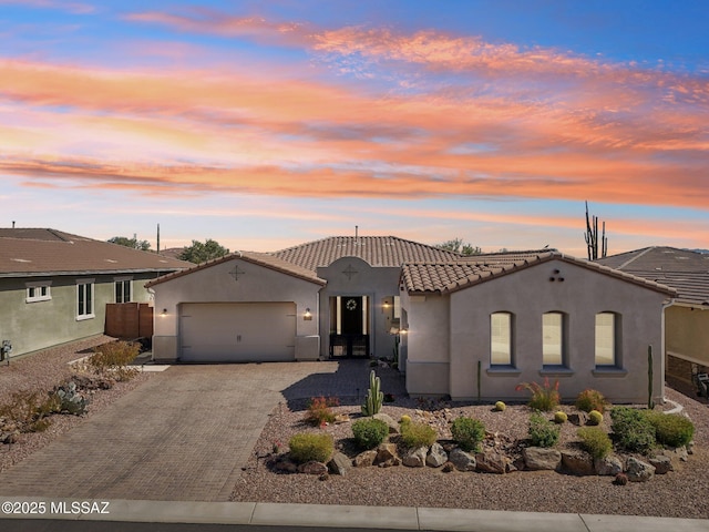 mediterranean / spanish-style home with an attached garage, a tile roof, fence, stucco siding, and decorative driveway