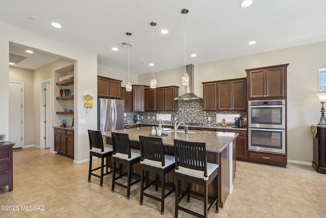 kitchen featuring visible vents, wall chimney exhaust hood, appliances with stainless steel finishes, an island with sink, and light stone counters