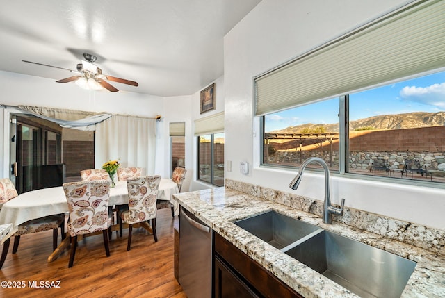 kitchen with a sink, a ceiling fan, wood finished floors, stainless steel dishwasher, and light stone counters