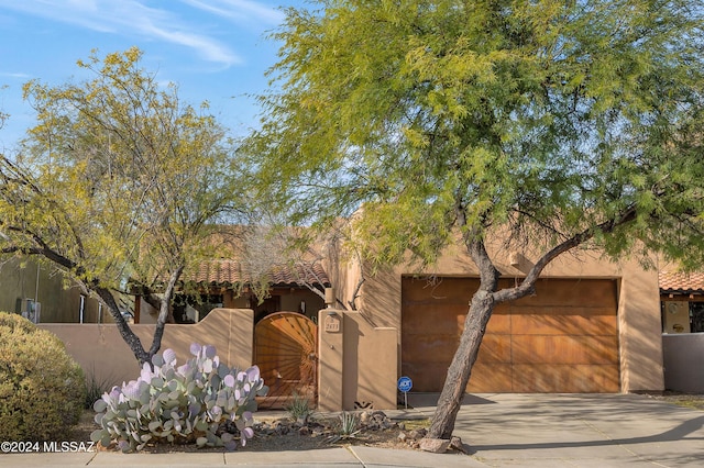 pueblo revival-style home with a tile roof, fence, an attached garage, driveway, and a gate