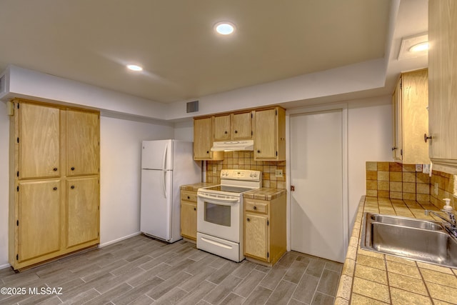 kitchen featuring a sink, tile countertops, under cabinet range hood, white appliances, and decorative backsplash