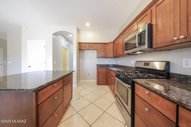 kitchen featuring stainless steel appliances, a center island, arched walkways, dark stone countertops, and brown cabinetry