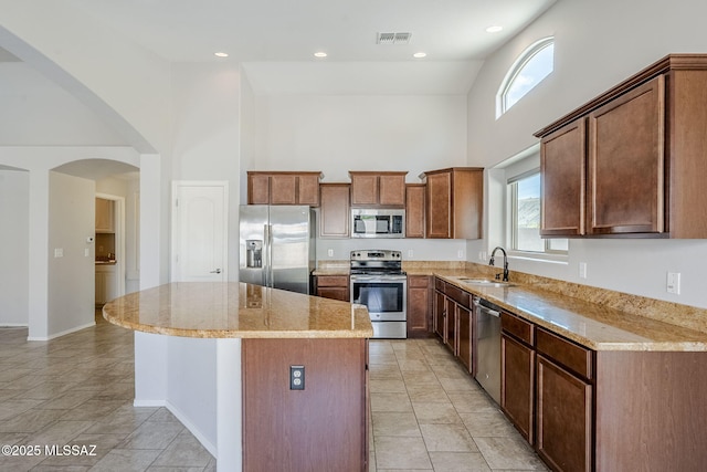 kitchen with a high ceiling, stainless steel appliances, a sink, light stone countertops, and a center island