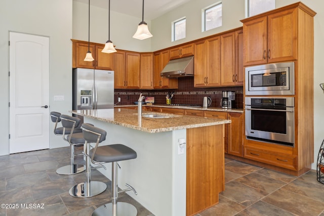 kitchen featuring decorative backsplash, under cabinet range hood, brown cabinets, and appliances with stainless steel finishes