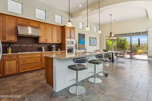 kitchen featuring brown cabinetry, stainless steel appliances, under cabinet range hood, light stone counters, and backsplash