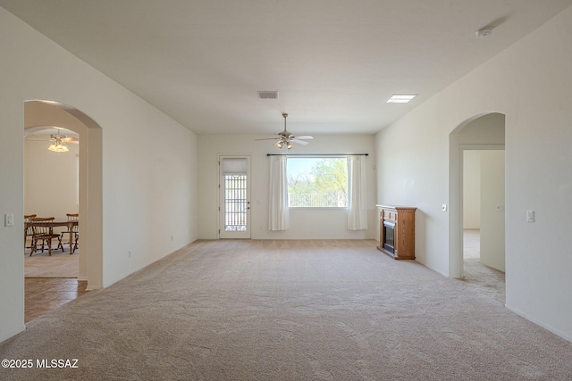 empty room with ceiling fan, visible vents, light colored carpet, arched walkways, and a glass covered fireplace