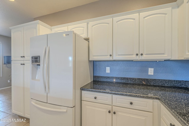 kitchen with light tile patterned flooring, dark stone countertops, white cabinets, and white refrigerator with ice dispenser