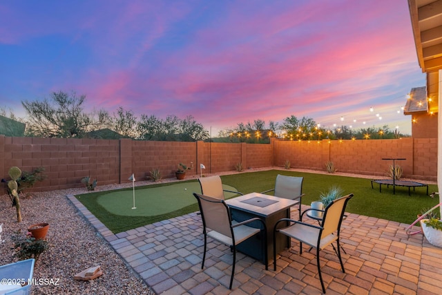 patio terrace at dusk with a fenced backyard, outdoor dining space, a lawn, and a trampoline