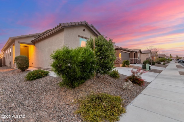 property exterior at dusk with stucco siding, an attached garage, a tiled roof, and driveway