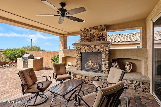 view of patio featuring an outdoor stone fireplace, fence, ceiling fan, a grill, and an outdoor kitchen