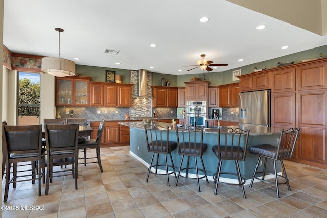 kitchen with recessed lighting, decorative backsplash, brown cabinetry, appliances with stainless steel finishes, and wall chimney exhaust hood