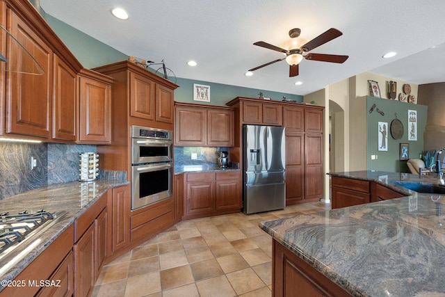 kitchen featuring arched walkways, decorative backsplash, appliances with stainless steel finishes, brown cabinets, and a ceiling fan