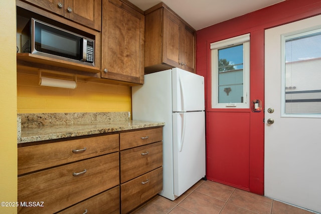 kitchen featuring stainless steel microwave, light tile patterned floors, brown cabinetry, and freestanding refrigerator