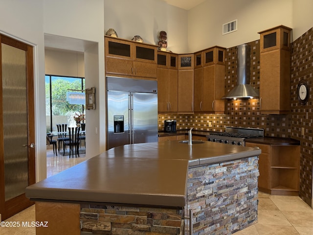 kitchen featuring stove, an island with sink, stainless steel built in refrigerator, visible vents, and wall chimney exhaust hood