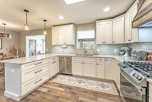 kitchen with a sink, wall chimney range hood, light wood-style flooring, a peninsula, and appliances with stainless steel finishes