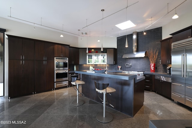 kitchen with wall chimney range hood, dark countertops, appliances with stainless steel finishes, a breakfast bar area, and decorative backsplash