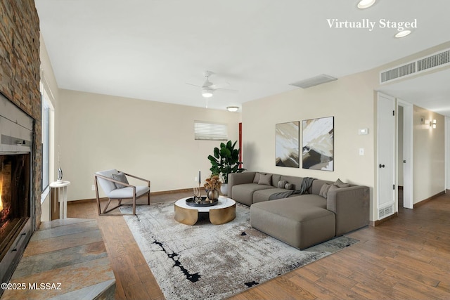 living room featuring baseboards, a warm lit fireplace, visible vents, ceiling fan, and wood finished floors